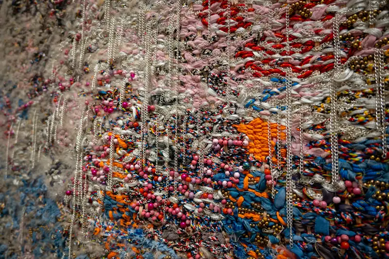 Nahaufnahme einer Textilarbeit aus Perlen, Fäden und Ketten in Rot, Blau und Orange