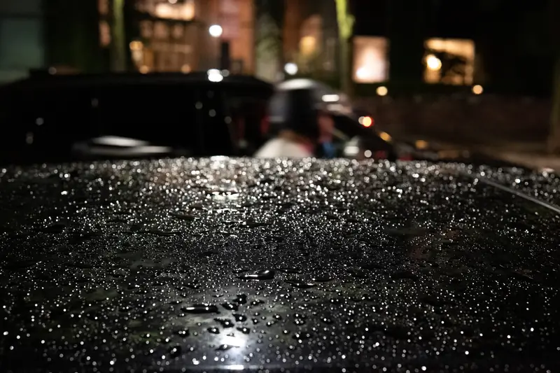 Regentropfen auf einem dunklen Autodach, dahinter die Lichter der Stadt
